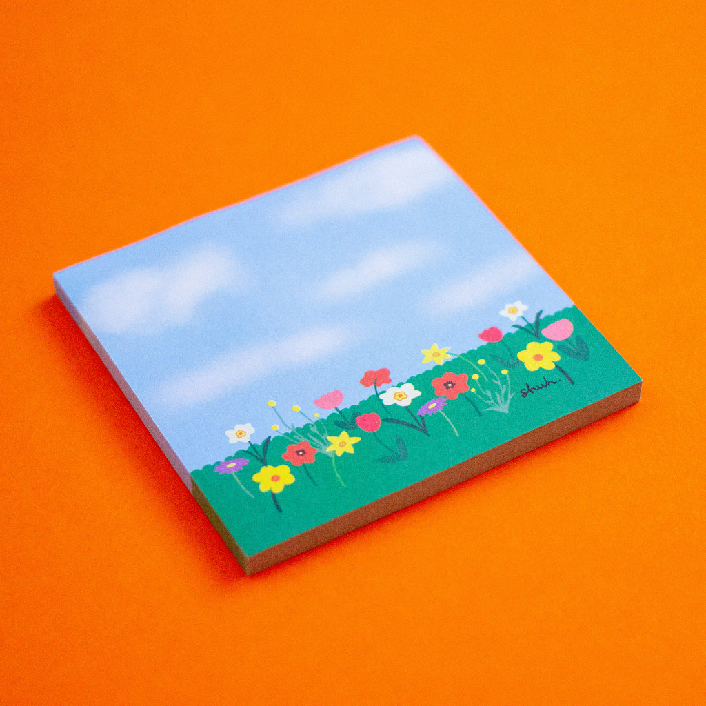 A square sticky note featuring a painted landscape of a blue sky with clouds and a green field of colourful wildflowers, displayed on a solid orange background.