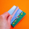 A hand holds a small sticky note with a colourful floral landscape illustration against a bright orange background.