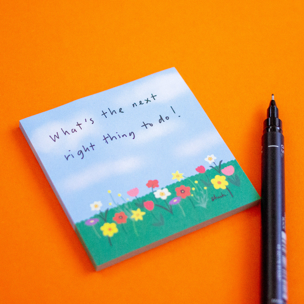 A square sticky note pad with a hand-painted sky and flower field design, featuring the handwritten quote 'What's the next right thing to do!', shown on an orange background next to a black pen.