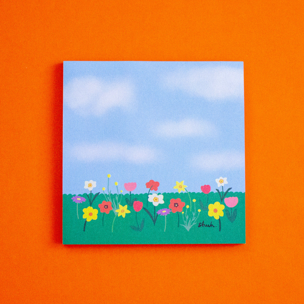 A square art print depicting a colourful flower meadow under a blue sky, displayed against a solid orange background.