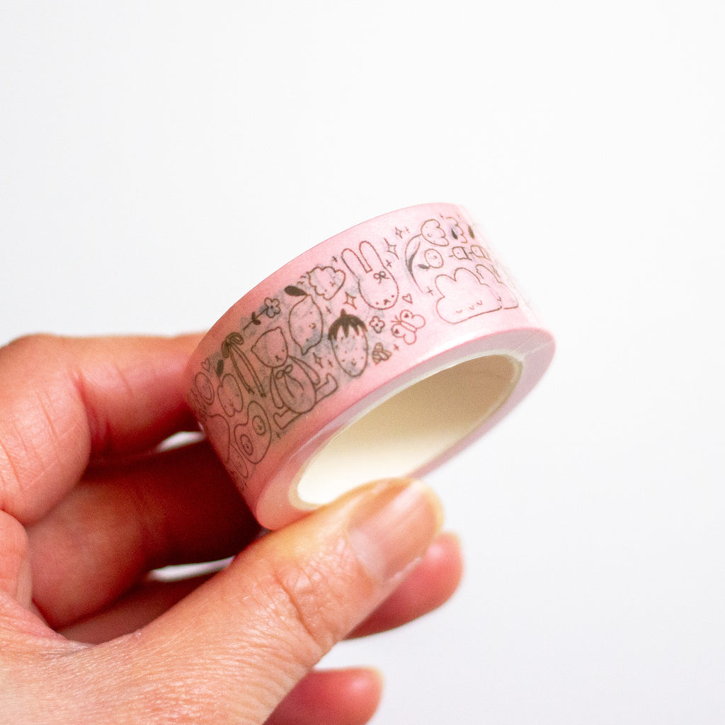 A hand holding a roll of light pink washi tape with a black cartoon pattern of strawberries, bunnies, and clouds against a plain white background.