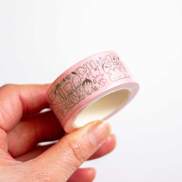 A hand holding a roll of light pink washi tape with a black cartoon pattern of strawberries, bunnies, and clouds against a plain white background.