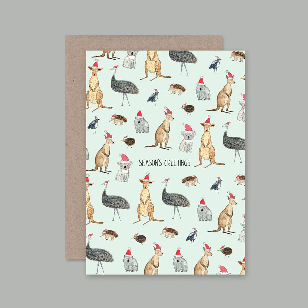 A greeting card with a light mint background and a repeating illustration pattern of kangaroos, emus, koalas, and birds, wearing red Santa hats, with the text 'SEASONS GREETINGS' in the center. The card is shown with a brown kraft envelope behind it on a solid grey background.