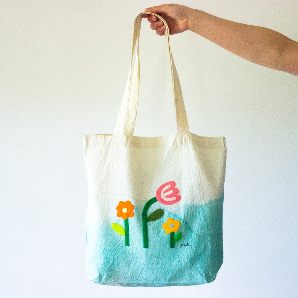 A hand holding the See You felt art tote bag. 2 daisies and a tulip, handstitched, on a died background. 