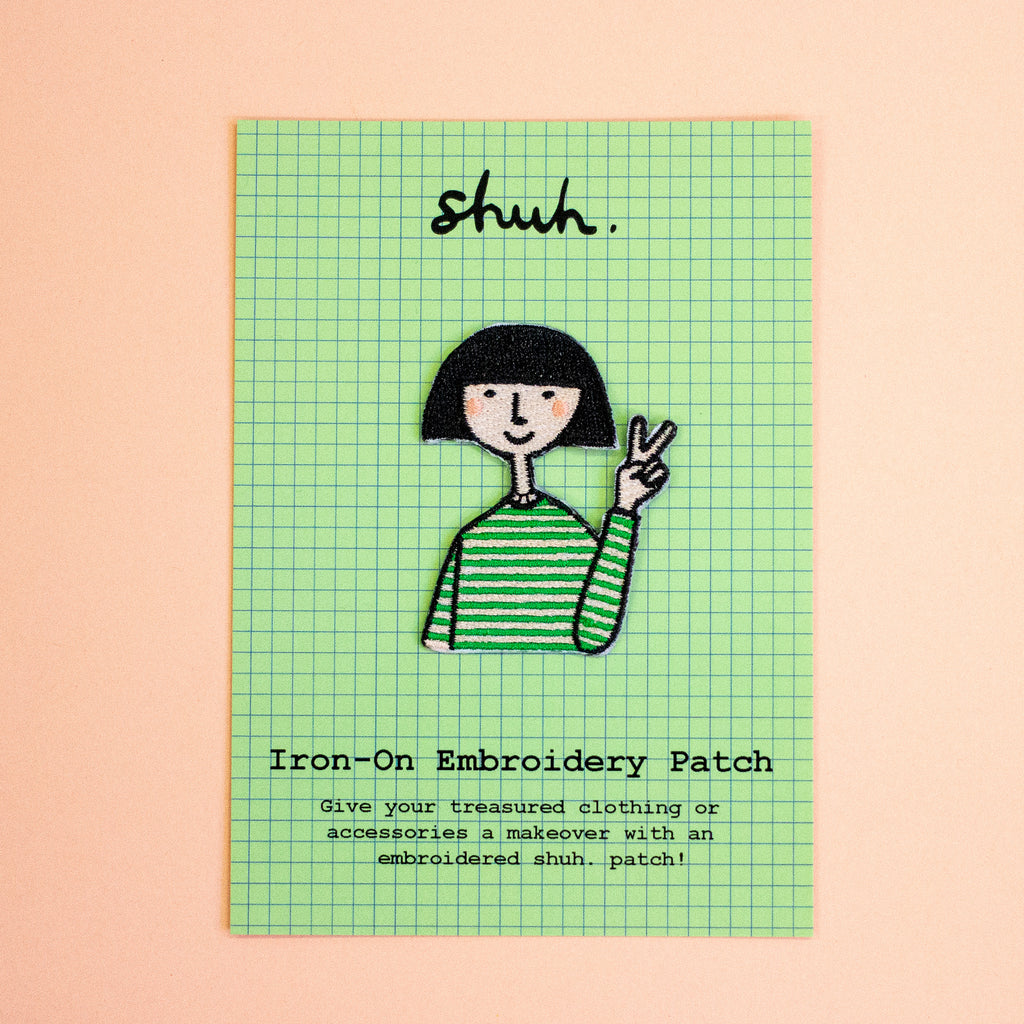 An iron-on embroidery patch shaped like a cartoon girl with a black bob haircut, a green and white striped shirt, and apeace hand sign on a green graph paper card with the word 'shuh.' at the top and text reading 'Iron-On Embroidery Patch' on a peach background.
