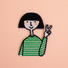 An embroidered patch of a cartoon girl with a black bob haircut, a green and white striped shirt, and a peace sign, set against a solid peach-pink background.