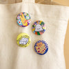 Four colourful round pin-back buttons displayed on a beige canvas tote bag. The buttons feature whimsical illustrations: a yellow bunny with flowers, a white cloud with the words 'I CAN DO IT', a yellow box labeled 'THOUGHTFUL TOFU' with a small smiley face, and cat holding a 'DINNER IDEAS' book. 