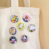 Six colourful round pin-back buttons with cute cartoon illustrations are arranged on a cream canvas tote bag against a warm beige background.