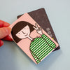 A hand holds a small illustrated card or sticker featuring a cartoon girl with dark hair, a pink grid background, and green-and-white striped clothing, with a second card partially visible behind it on a pale blue surface.
