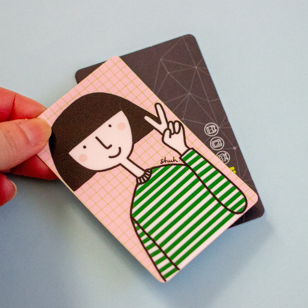 A hand holds a small illustrated card or sticker featuring a cartoon girl with dark hair, a pink grid background, and green-and-white striped clothing, with a second card partially visible behind it on a pale blue surface.