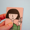 A hand holding a square card featuring a minimalist cartoon illustration of a girl with a black bob haircut, pink grid background, and green-and-white striped top, against a pale blue background.