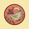 A circular embroidered patch featuring a white goose wearing a blue bow, with the words 'SILLY GOOSE' in black text, set against a dusty pink background with a dark red stitched border, shown on a pale yellow background.