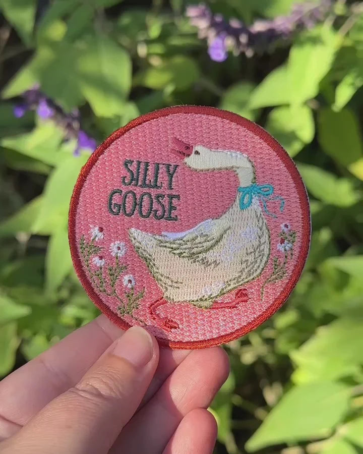 A hand holds a round embroidered patch featuring a white goose with a blue bow, pink background, and the words 'SILLY GOOSE' against a blurred green garden background.