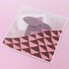 An illustrated square print of a rat sleeping on a pillow and tucked in under a blanket with white border photographed against a pink backdrop. 