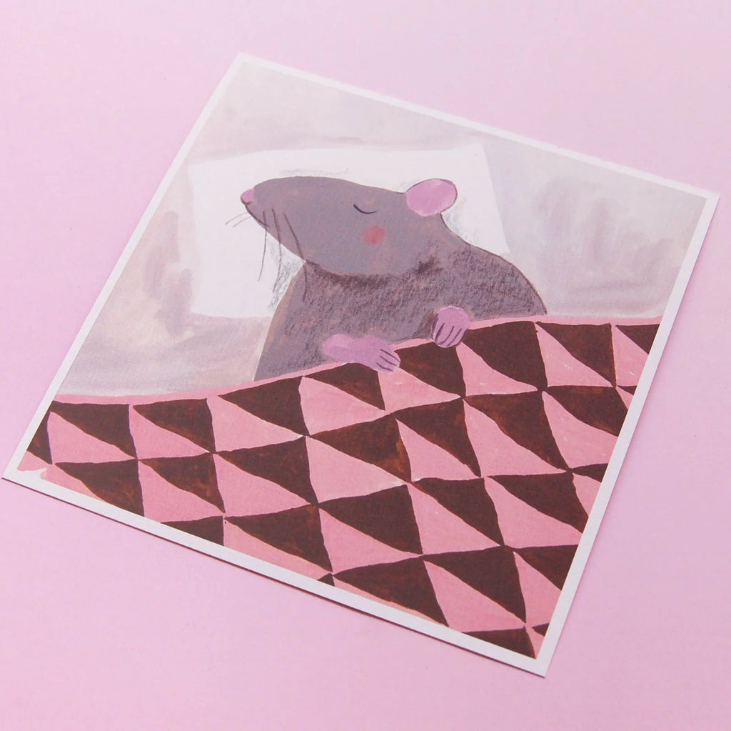 An illustrated square print of a rat sleeping on a pillow and tucked in under a blanket with white border photographed against a pink backdrop. 