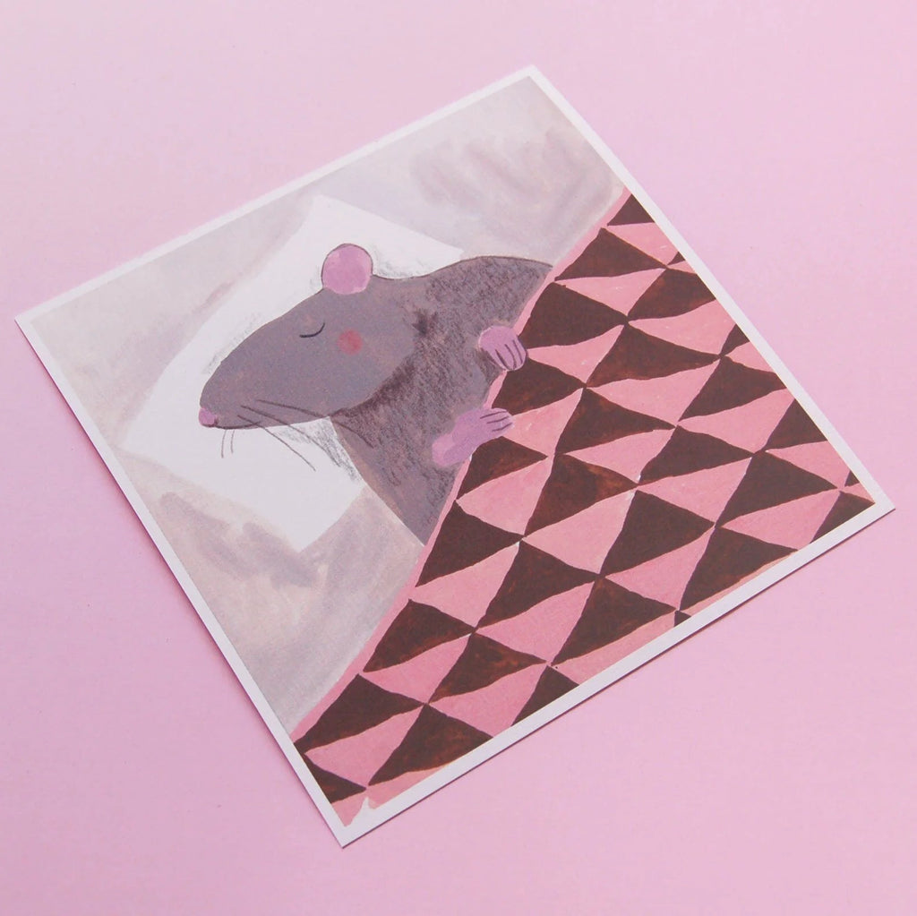 An illustrated square print of a rat sleeping on a pillow and tucked in under a blanket with white border photographed against a pink backdrop. 