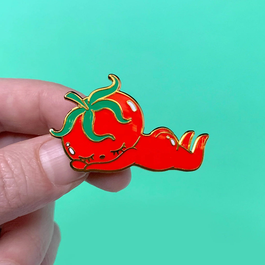 A hand holding a small enamel pin shaped like a stylized red tomato with green leaves and a long red tail, set against a solid mint green background.