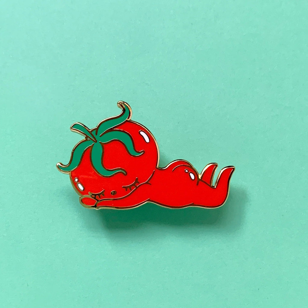 A red tomato-shaped enamel pin with green stem details, resting on a solid mint green background.
