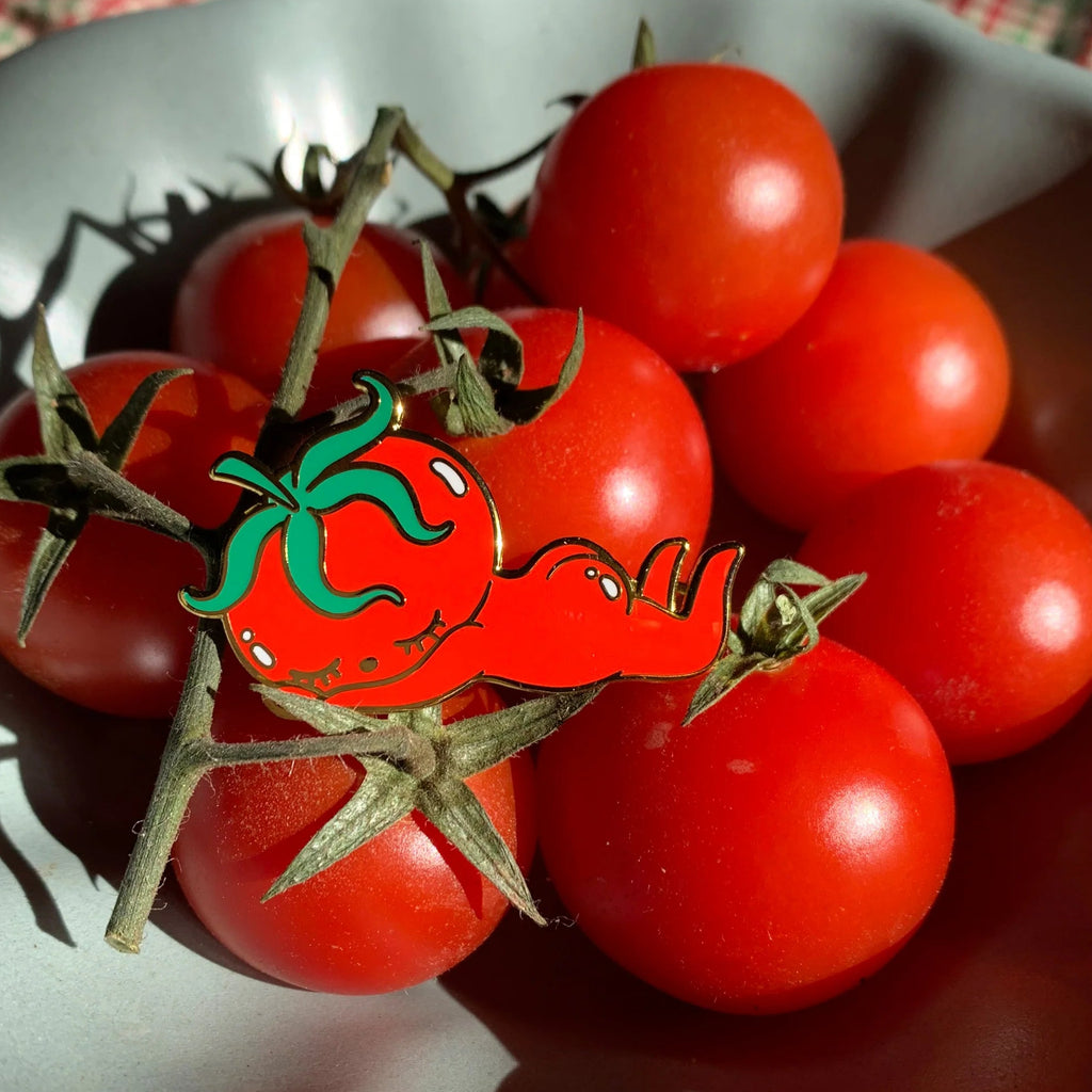 A red tomato-shaped enamel pin with green leaves, pinned to a vine of fresh red cherry tomatoes in a grey ceramic bowl.