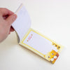 A hand holding open a small notepad with a cute cartoon cat illustration on the cover. The notepad has a yellow border and a white writing area, photographed on a plain white background.