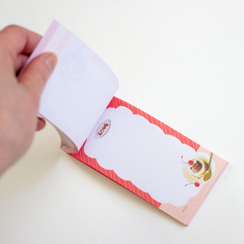 A hand holding open a small notepad with a pink and red striped cover and a large blank writing area, photographed on a plain light background.