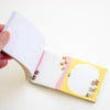 A hand holding open a small notepad with white pages. The cover is yellow with small illustrated characters and a pink gingham stripe, photographed on a clean white background.