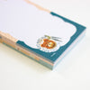 A close-up of a stack of notepad sheets with a teal border and a printed food illustration near the bottom, photographed on a clean white background.