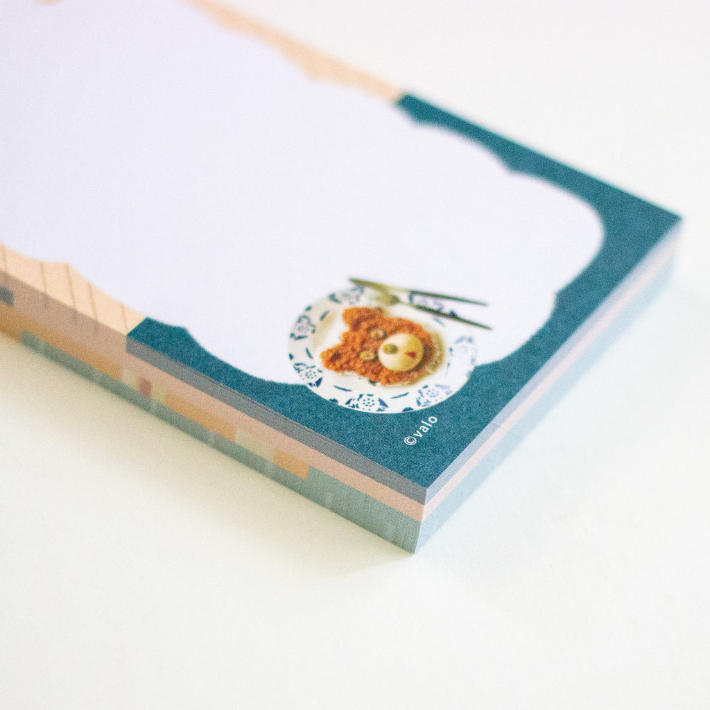 A close-up of a stack of notepad sheets with a teal border and a printed food illustration near the bottom, photographed on a clean white background.