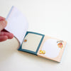 A hand holding open a small pastel notepad with cute cartoon illustrations and two lined writing sections on a clean white background.