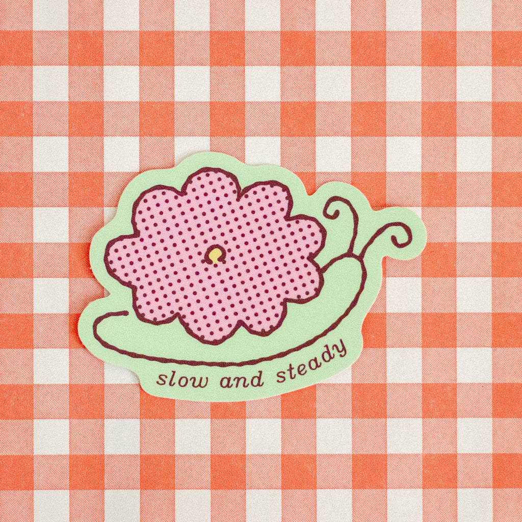 A die-cut sticker featuring a pink flower with a dotted print on a light green snail-like shape, with the words 'slow and steady' in brown script. The sticker is placed on a red and white gingham checkered background.