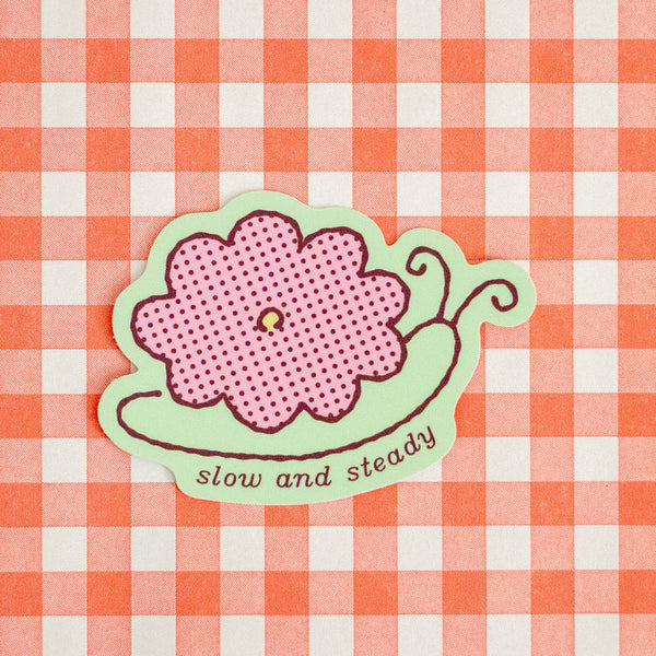 A die-cut sticker featuring a pink flower with a dotted print on a light green snail-like shape, with the words 'slow and steady' in brown script. The sticker is placed on a red and white gingham checkered background.