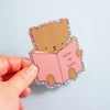 A hand holding a die-cut sticker featuring a cute brown cartoon bear reading a pink book with the text 'HOW TO SLAY' on a light blue background.