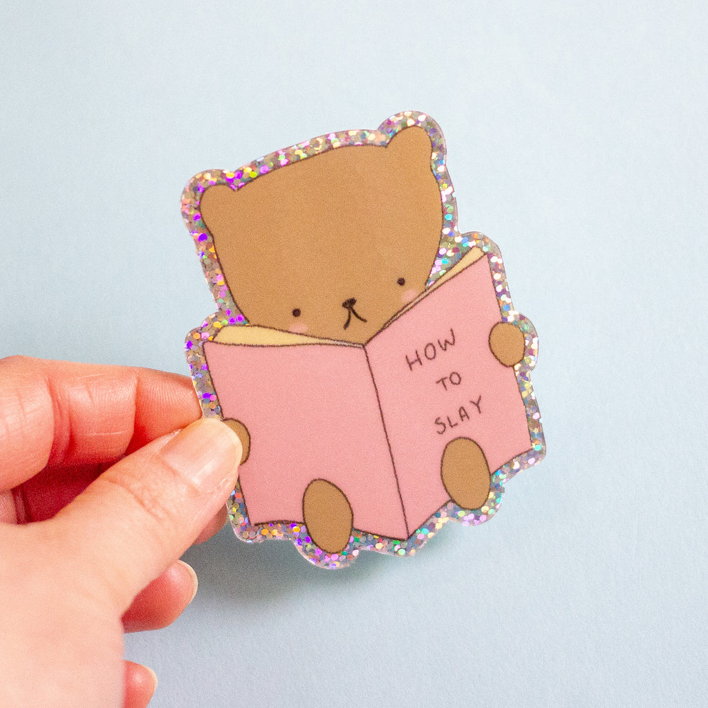 A hand holding a die-cut sticker featuring a cute brown cartoon bear reading a pink book with the text 'HOW TO SLAY' on a light blue background.