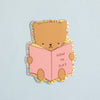 A cute cartoon bear sticker reading a pink book that says 'HOW TO SLAY' on a light blue background.