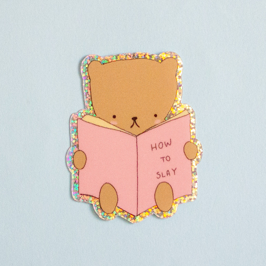 A cute cartoon bear sticker reading a pink book that says 'HOW TO SLAY' on a light blue background.