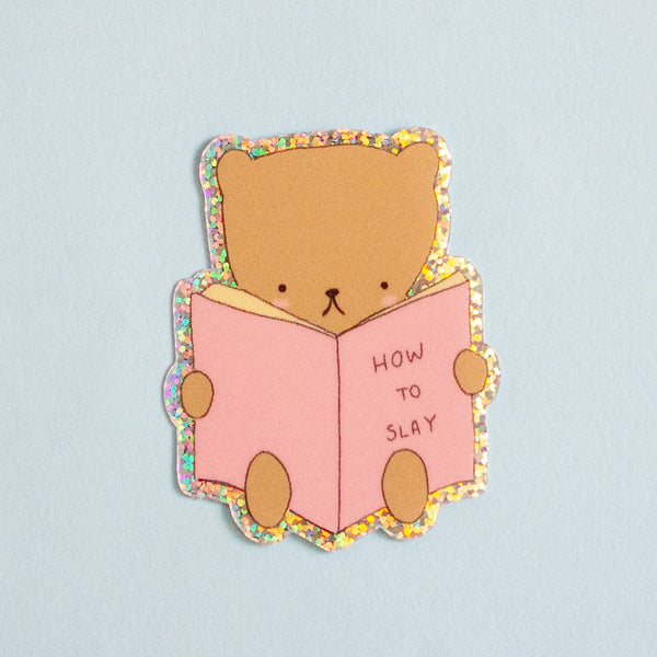A cute cartoon bear sticker reading a pink book that says 'HOW TO SLAY' on a light blue background.