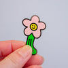 A small enamel pin shaped like a smiling flower with a pink bloom, yellow center, and green stem, held between two fingers against a plain light grey background.