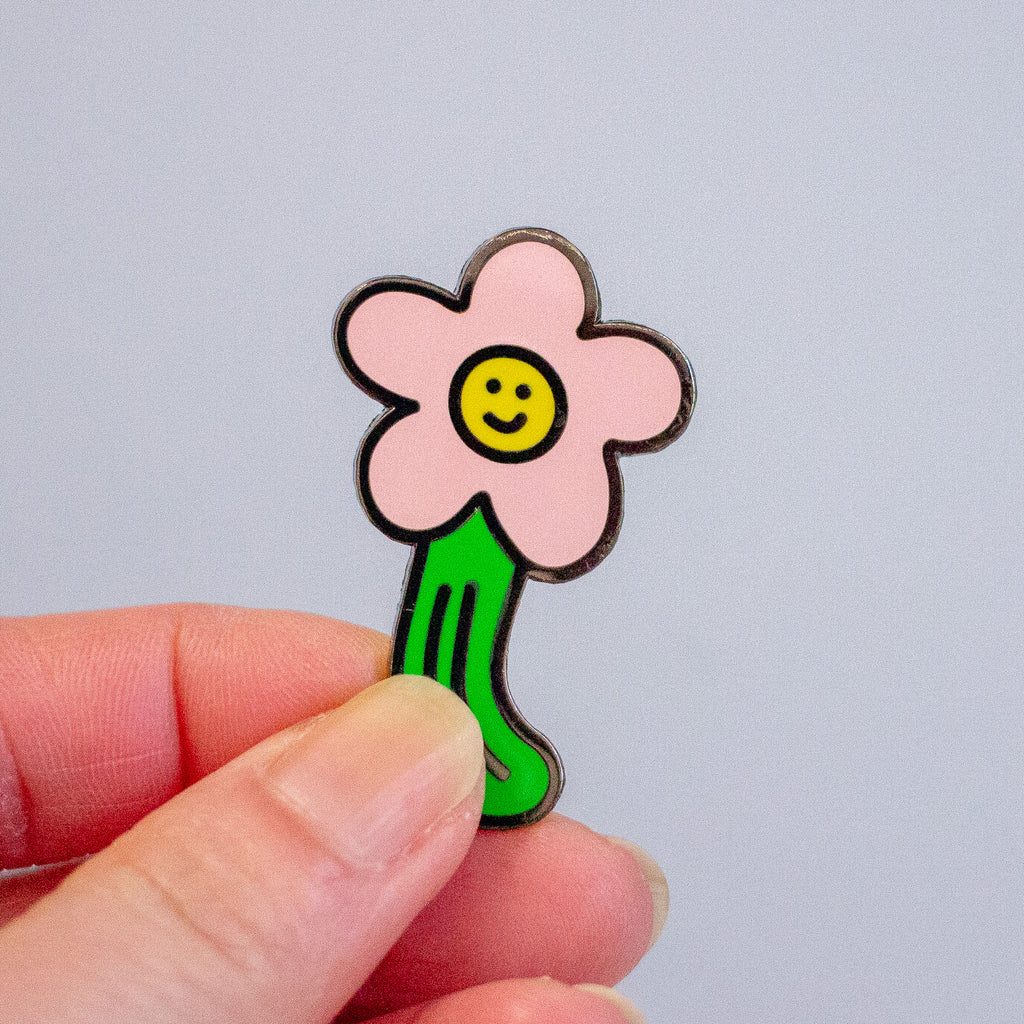 A small enamel pin shaped like a smiling flower with a pink bloom, yellow center, and green stem, held between two fingers against a plain light grey background.