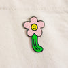 A small enamel pin shaped like a flower with a pink bloom, yellow smiling face center, and green stem, placed on a light fabric background.