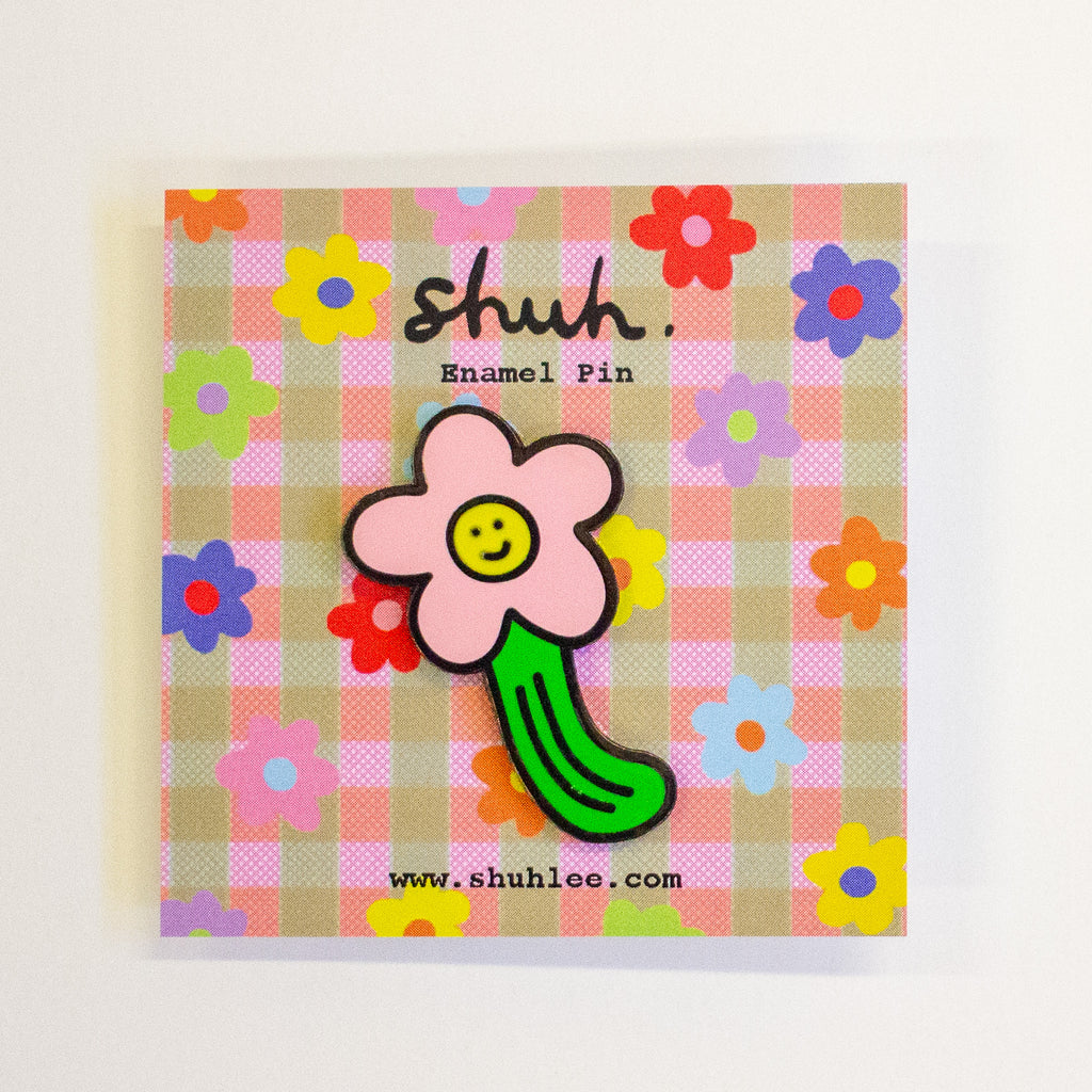 A enamel pin shaped like a pink flower with a green stem and a yellow smiley face, mounted on a pink and beige plaid backing card with colourful floral patterns and the brand name 'shuh.'.
