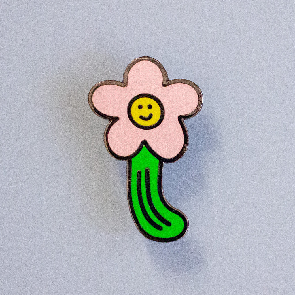 A flower-shaped enamel pin with a light pink five-petal bloom, a yellow smiley face center, and a bright green stem, shown on a plain light grey background.