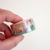 A hand holding a roll of washi tape featuring a colourful illustration of cat figurines with gold glitter dots, against a plain off-white background.