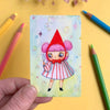 Colourful illustration of a character on a card held by a hand with coloured pencils around.