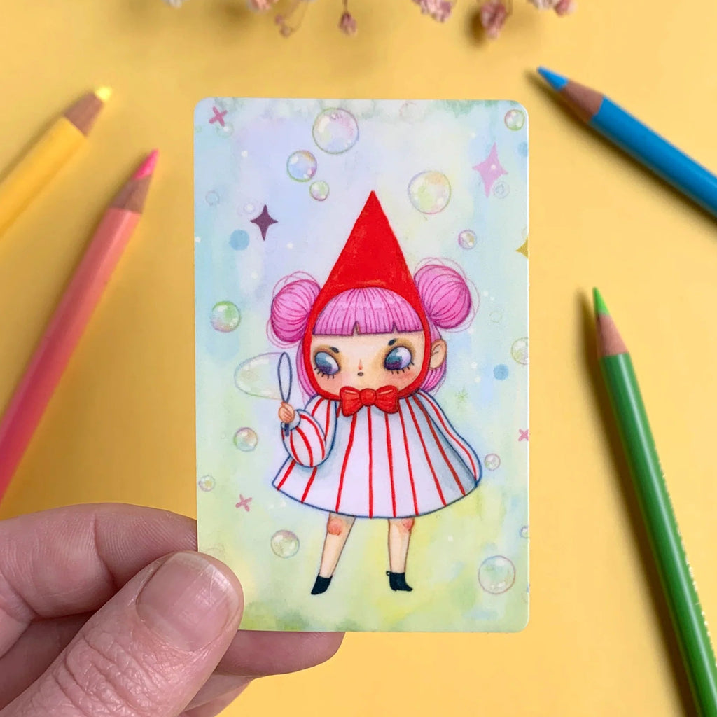 Colourful illustration of a character on a card held by a hand with coloured pencils around.