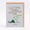 A light blue greeting card with a kraft envelope behind it, featuring a cartoon green insect-like character lying on the ground and the text 'DAD YOU'RE DOING A SPECTACULAR JOB' and 'I MEAN, LOOK AT ME.'.