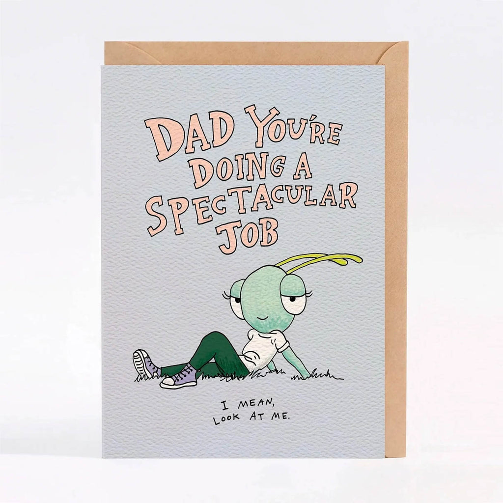 A light blue greeting card with a kraft envelope behind it, featuring a cartoon green insect-like character lying on the ground and the text 'DAD YOU'RE DOING A SPECTACULAR JOB' and 'I MEAN, LOOK AT ME.'.