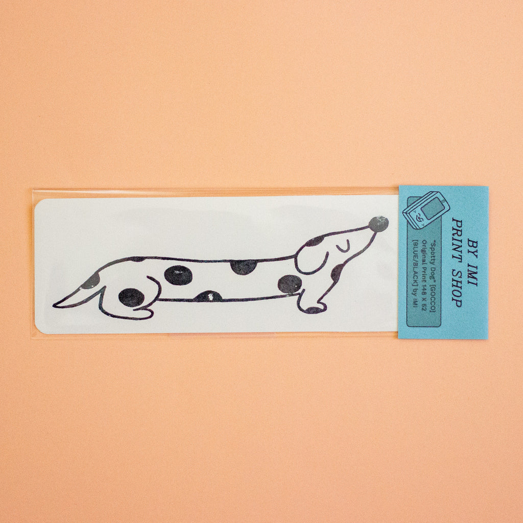 A clear plastic sleeve holds a white rectangular print featuring a black line-art dachshund with black spots, attached to a light blue header card that reads 'BY IMI PRINT SHOP.' The product is photographed against a solid peach background.