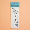 A clear plastic sleeve holds a white rectangular print featuring a black line-art dachshund with black spots, attached to a light blue header card that reads 'BY IMI PRINT SHOP.' The product is photographed against a solid peach background.