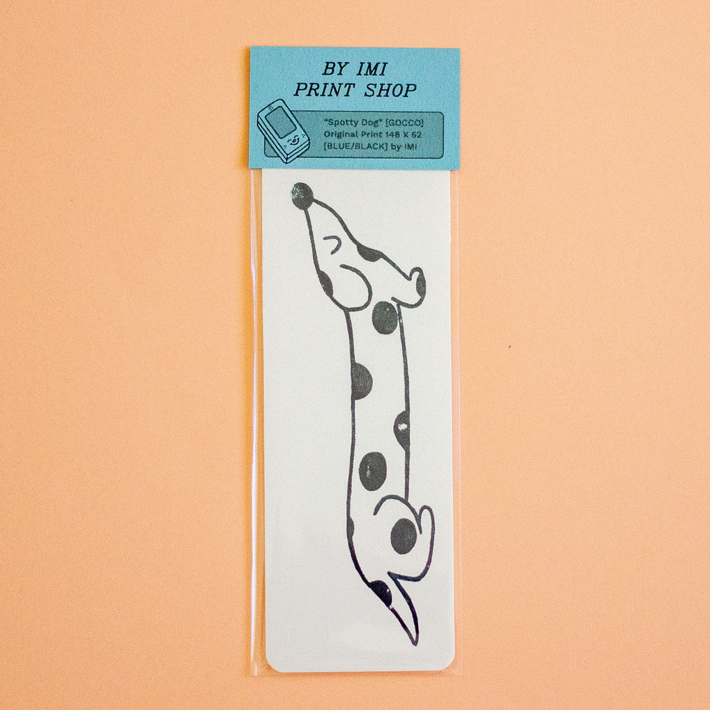 A clear plastic sleeve holds a white rectangular print featuring a black line-art dachshund with black spots, attached to a light blue header card that reads 'BY IMI PRINT SHOP.' The product is photographed against a solid peach background.