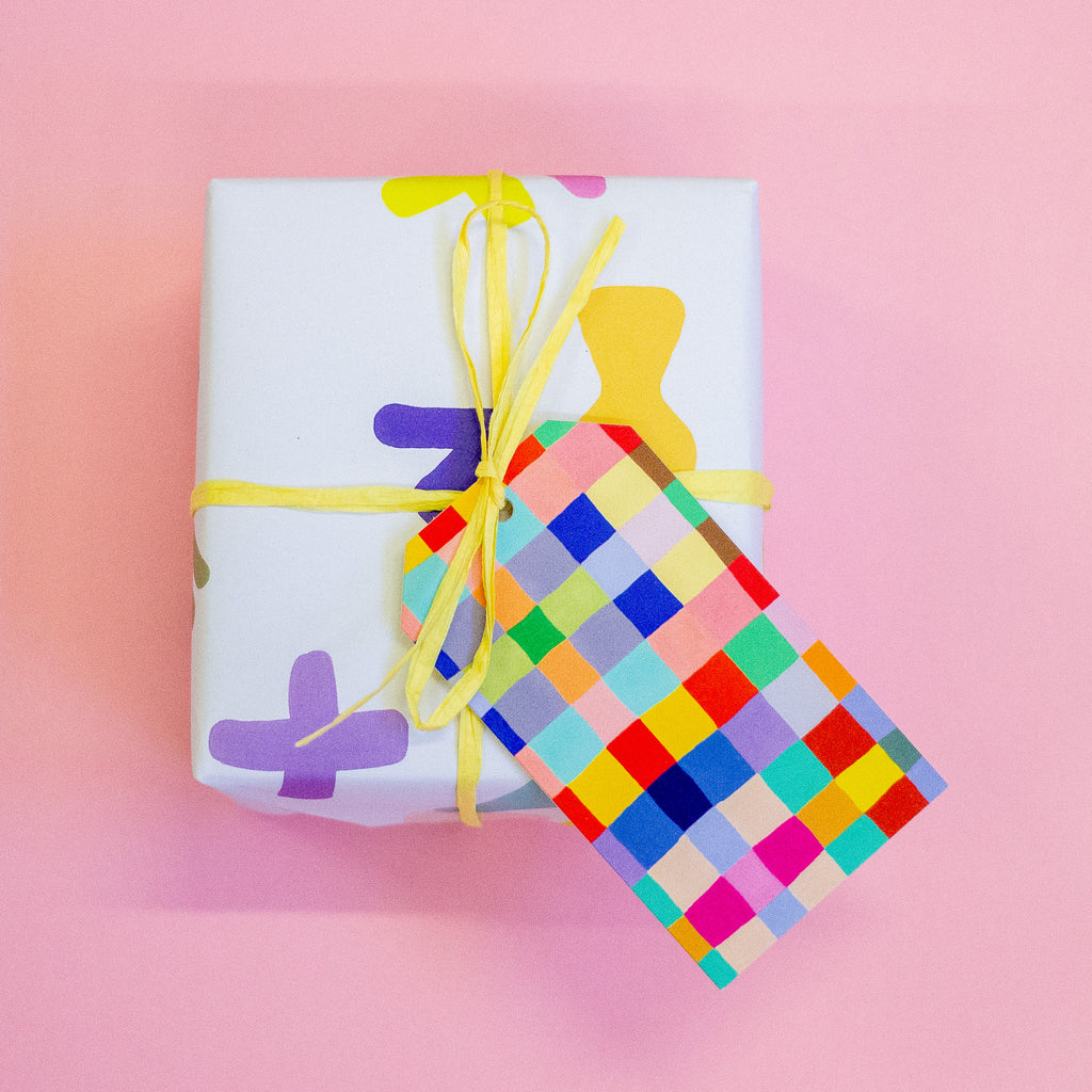 A closeup picture of the squares gift tag tied to a yellow string on a gift box. Print features multicoloured hand drawn squares, creating a vibrant chequered pattern. 